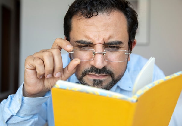 A man holding his eyeglasses and squinting while reading a book.<br />
